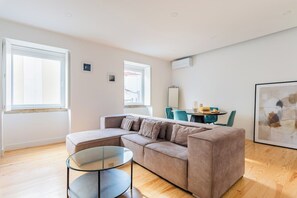 A modern living room with a sectional sofa, a glass coffee table, and a dining area with chairs and a table. -  2BR Chic&Bright com 120m2 em Campo de Ourique (Lisbon)