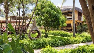 Explore a serene tropical garden with lush greenery, palm trees, and a well-maintained lawn, featuring a unique wicker structure and a thatched-roof building in the background.