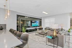 Una sala de estar moderna con un televisor de pantalla plana grande, un juego de sofá, una zona de comedor con una mesa larga y un espejo decorativo.
