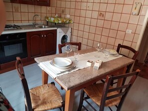 Comedor con una mesa dispuesta para cuatro, incluyendo platos, vasos y cubiertos. Hay dos sillas con asientos de mimbre. Se ve una cocina con estufa a gas y lavadora al fondo.