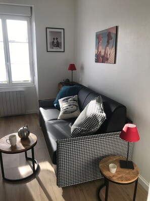 Un salon moderne avec un canapé en cuir noir, des coussins à motifs, une petite table ronde sur laquelle se trouvent une théière et une tasse, un lampadaire rouge et deux tableaux encadrés accrochés au mur.