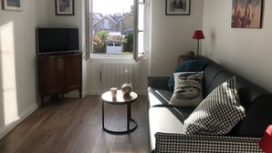 Un salón con sofá, televisor, una mesita redonda con una taza y una tetera, un radiador y una ventana que ofrece vistas a casas.
