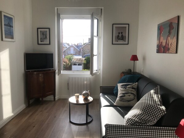 Un salon avec un canapé, un téléviseur, une petite table ronde sur laquelle se trouvent une tasse et une théière, un radiateur et une fenêtre donnant sur les maisons à l’extérieur.