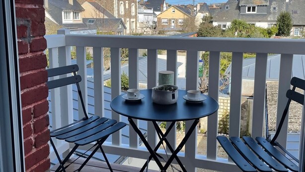 Una terraza de madera con una mesa redonda y dos sillas, con vista a una zona residencial con casas y un cielo despejado.