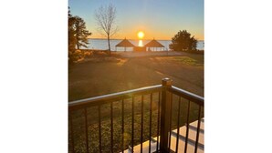 A sunset view from a balcony with a railing, overlooking a beach with two beach huts and a clear sky. - Coastal Farmhouse Getaway! (White Stone)