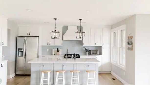A modern kitchen with a central island, bar stools, stainless steel appliances, and pendant lighting. - Coastal Farmhouse Getaway! (White Stone)