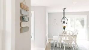 A modern dining area with a white table and chairs, a hanging light fixture, and a window with natural light. - Coastal Farmhouse Getaway! (White Stone)