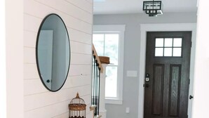 A hallway with a wooden console table, a round mirror, and a decorative birdcage. A patterned rug leads to a dark door. - Coastal Farmhouse Getaway! (White Stone)