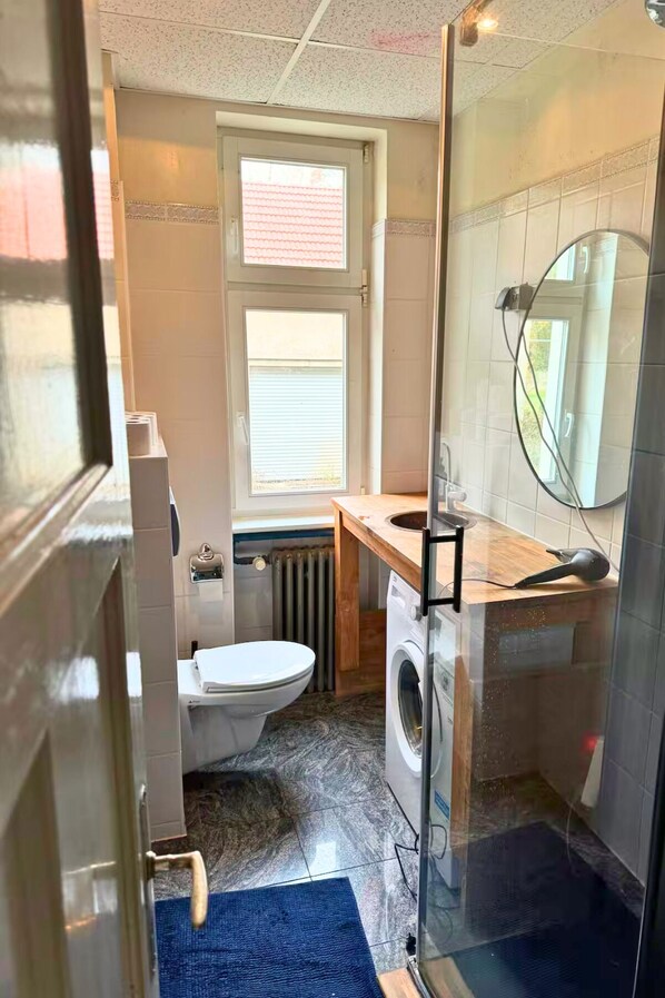 Casa de banho compacta com sanita, máquina de lavar roupa e cabine de duche em vidro.
