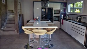 A modern kitchen with a central island, bar stools, a built-in oven, and a window with a view of greenery. - Private heated pool and jacuzzi villa (Furdenheim)