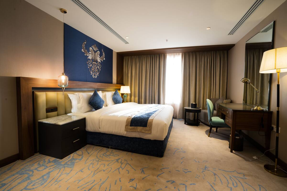 A hotel room with a large bed, bedside tables, a desk, a chair, a lamp, a mirror, and a decorative wall piece.