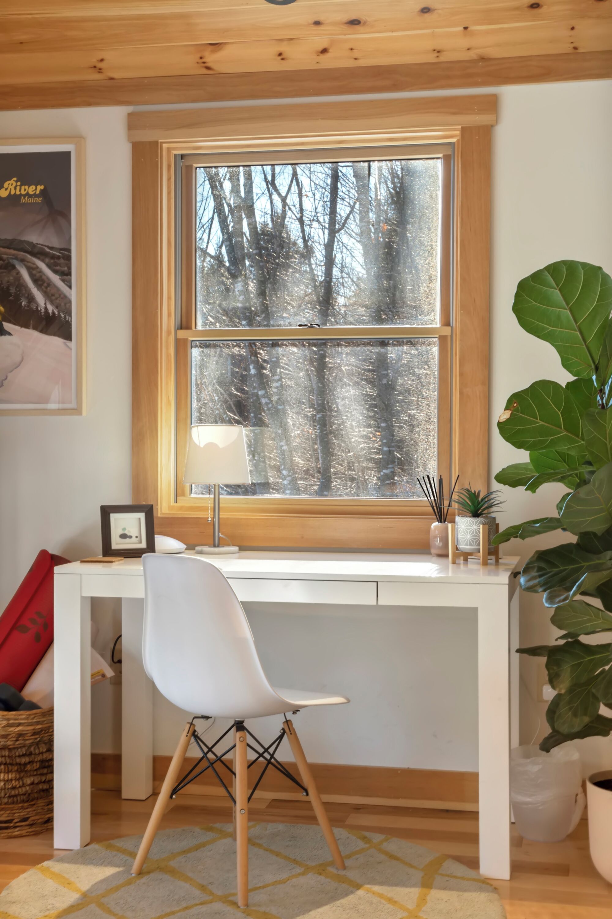 A room with a white desk, a chair, a large window with a view of trees, a lamp, and a plant.