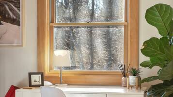 A room with a white desk, a chair, a large window with a view of trees, a lamp, and a plant.