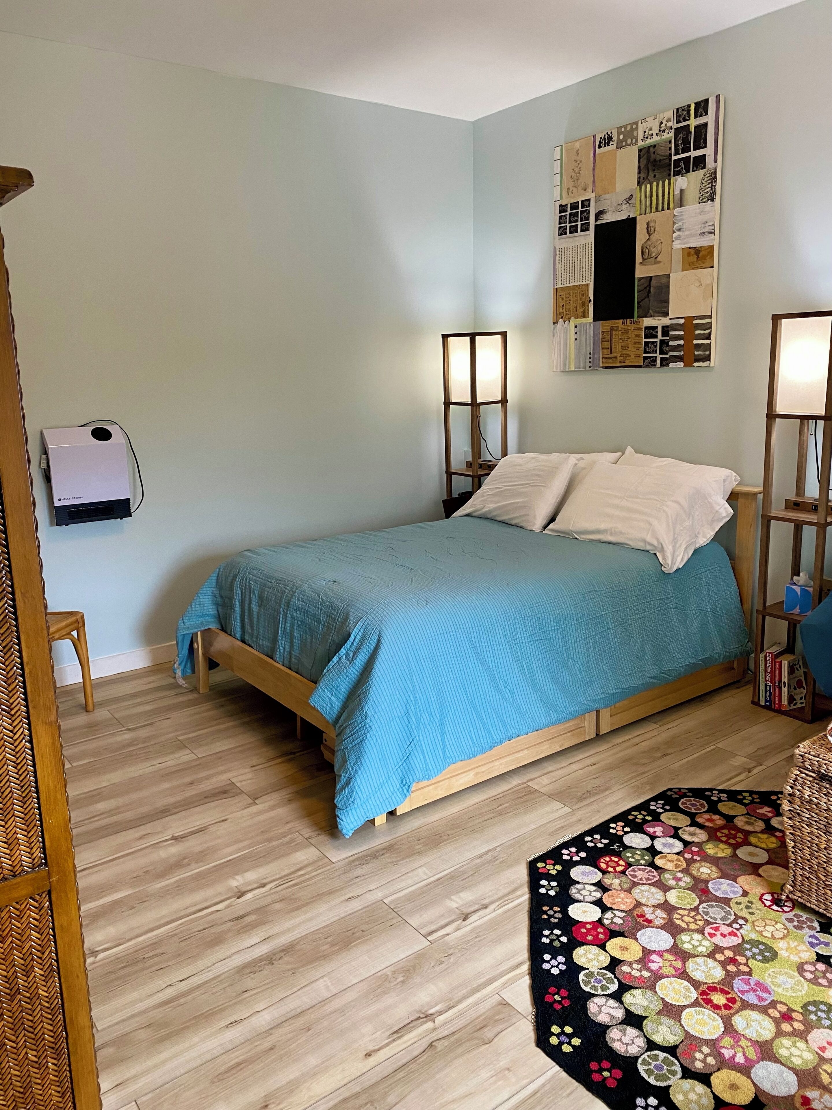 A bedroom with a bed, a standing lamp, a wall-mounted heater, and a decorative rug.