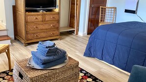 Une chambre avec un téléviseur posé sur un meuble en bois, un lit recouvert d’une couverture bleue, un pouf en osier avec des serviettes et un tapis coloré.
