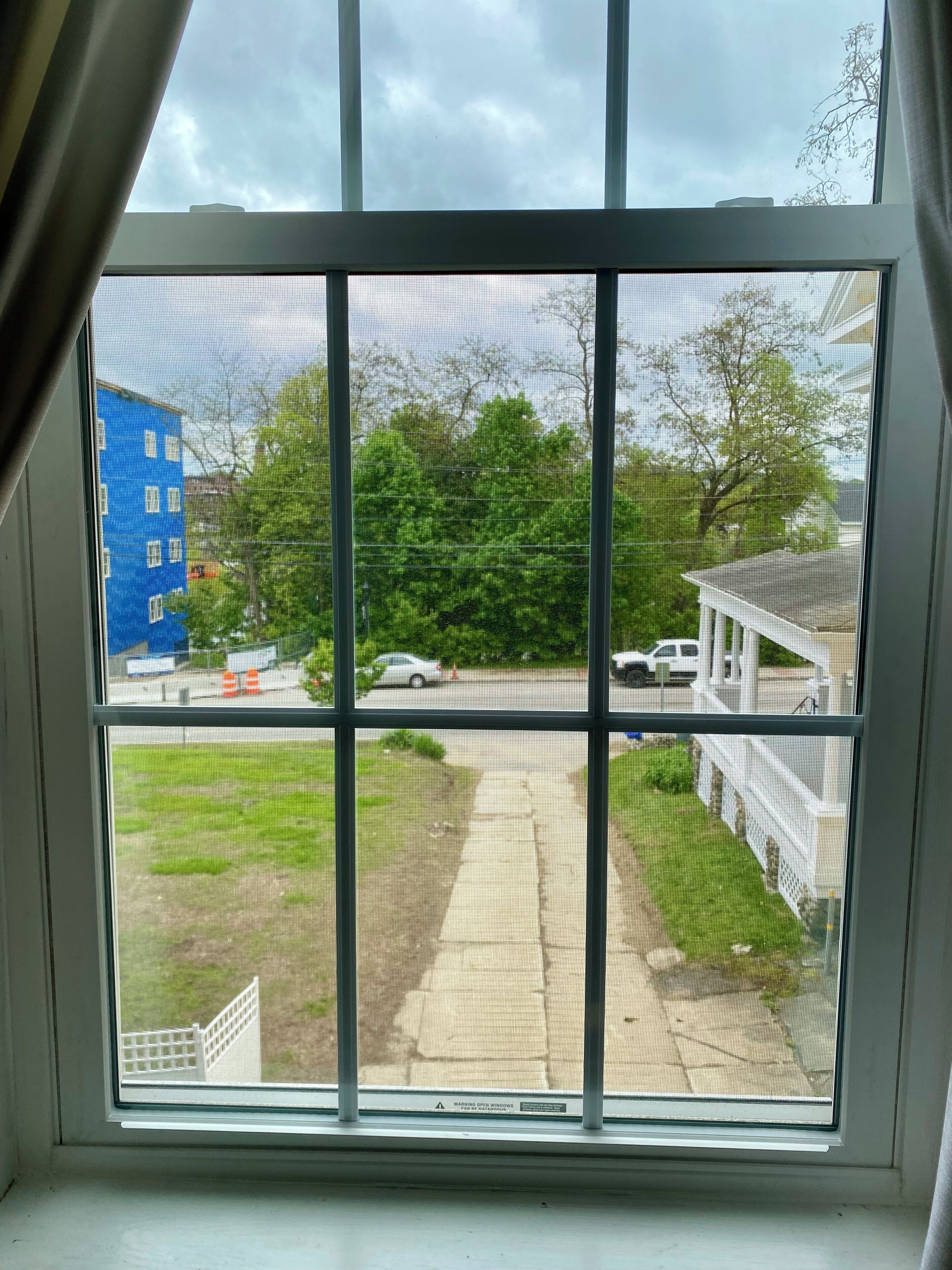Une vue à travers une fenêtre ornée d’une grille métallique, donnant sur une rue bordée d’immeubles, d’arbres et d’un trottoir.