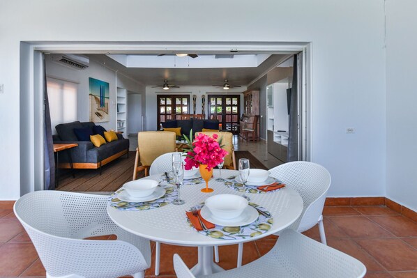 A dining area with a round table set for four, surrounded by white chairs. A vase with pink flowers is on the table, and there are plates, bowls, and cutlery.