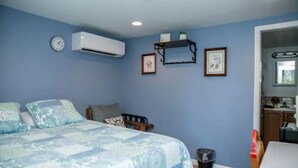 A bedroom with a bed, a clock, an air conditioner, a shelf with pictures, and a door leading to a kitchen.