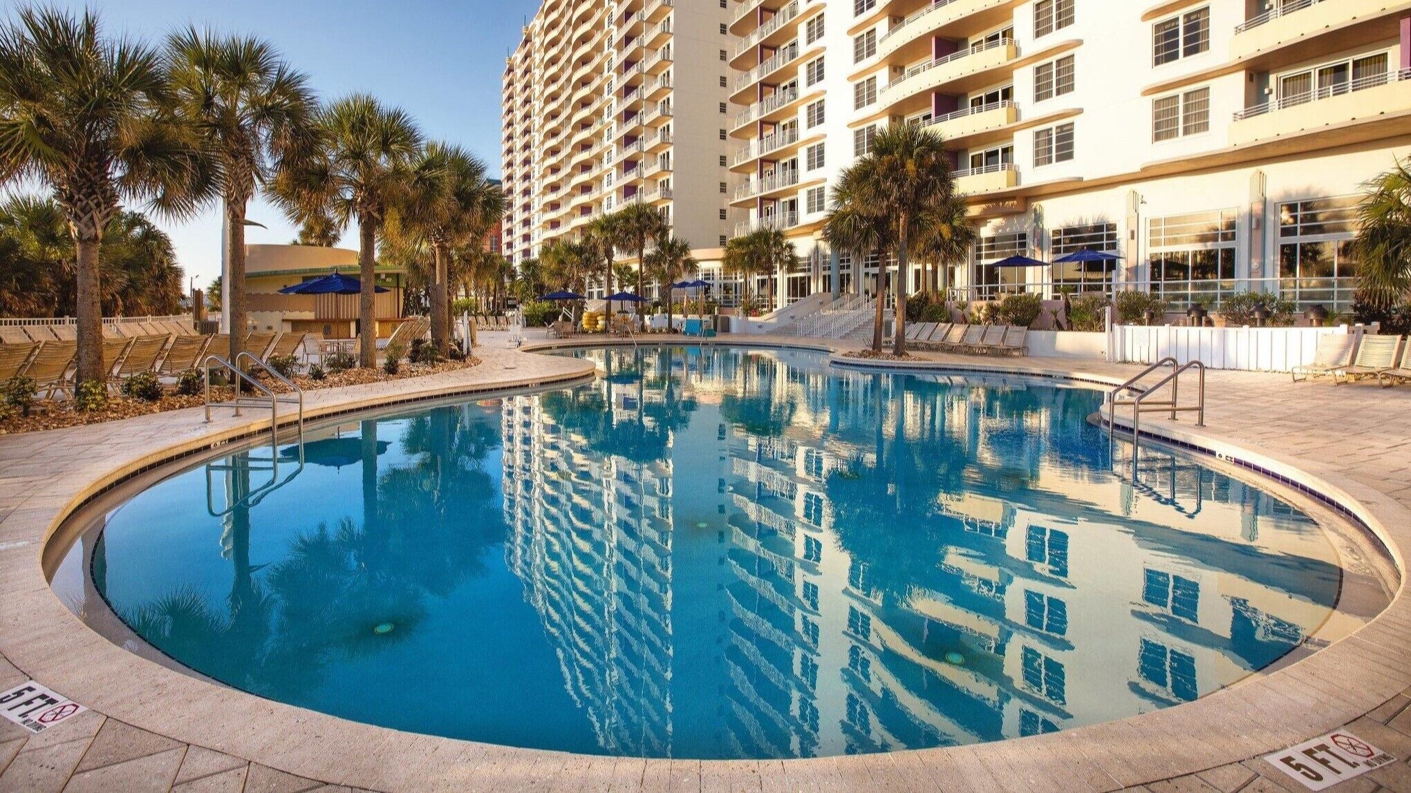 A swimming pool surrounded by palm trees and a multi-story hotel building.