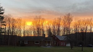 Een huis met voortuin en garage, omringd door bomen en een zonsondergang op de achtergrond.