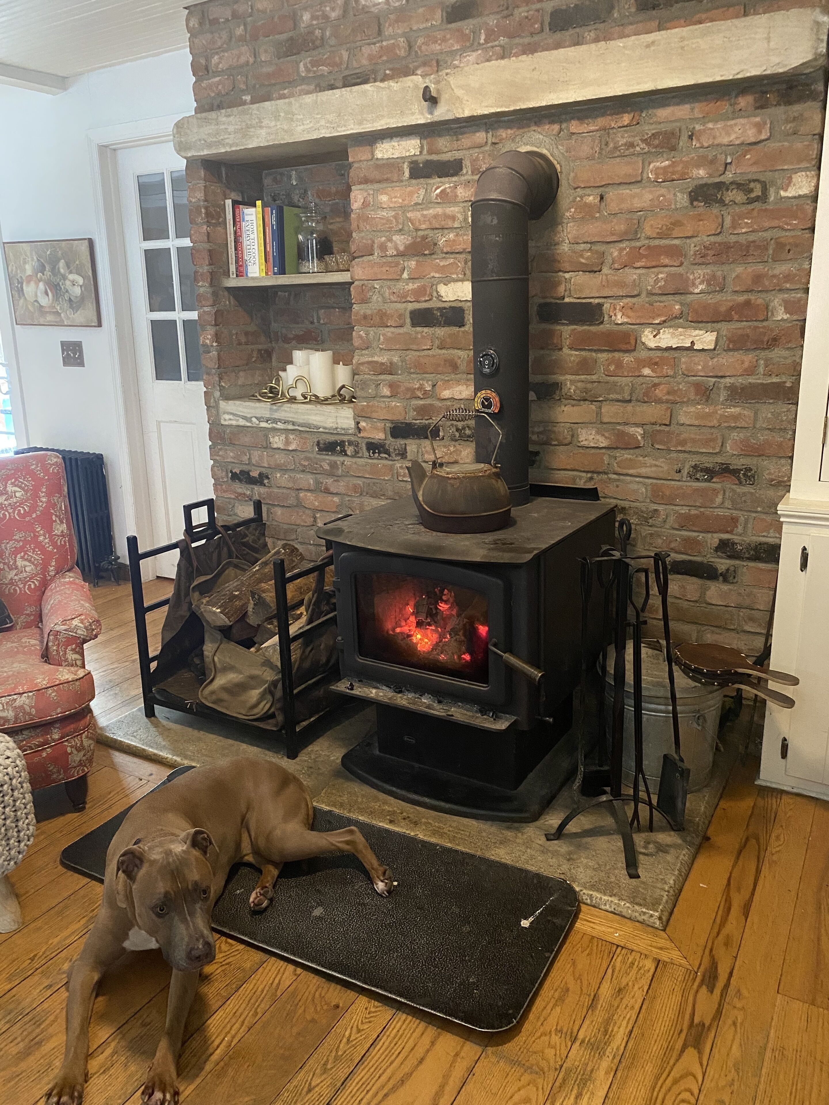 A room with a brick fireplace, a dog lying on a mat, and a red armchair.
