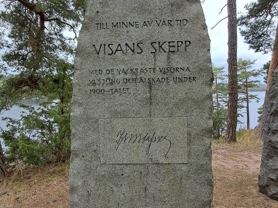A stone monument with inscriptions, surrounded by trees and a body of water.