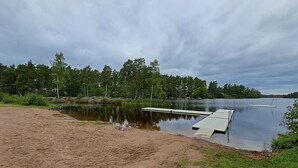 A lakeside area with a sandy beach, a wooden dock, and a calm lake surrounded by trees. - Ankis Bed and Breakfast (Storebro)