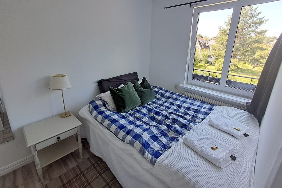Un dormitorio con cama, mesita de noche, lámpara y ventana con vistas a zonas verdes.