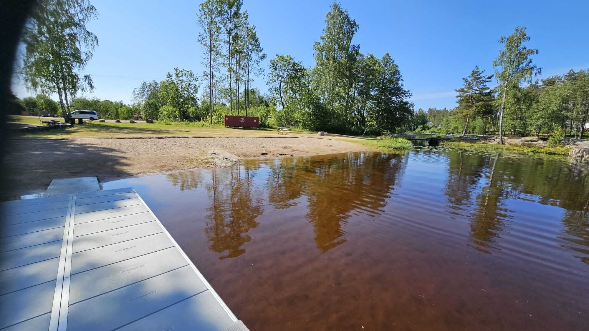 Una terraza de madera con vista a un cuerpo de agua tranquilo, una mesa de picnic y un contenedor rojo al fondo.