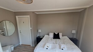 A hotel room with a large bed, two bedside tables with lamps, a round mirror on the wall, a white chair, and a door. - Ankis Bed and Breakfast (Storebro)