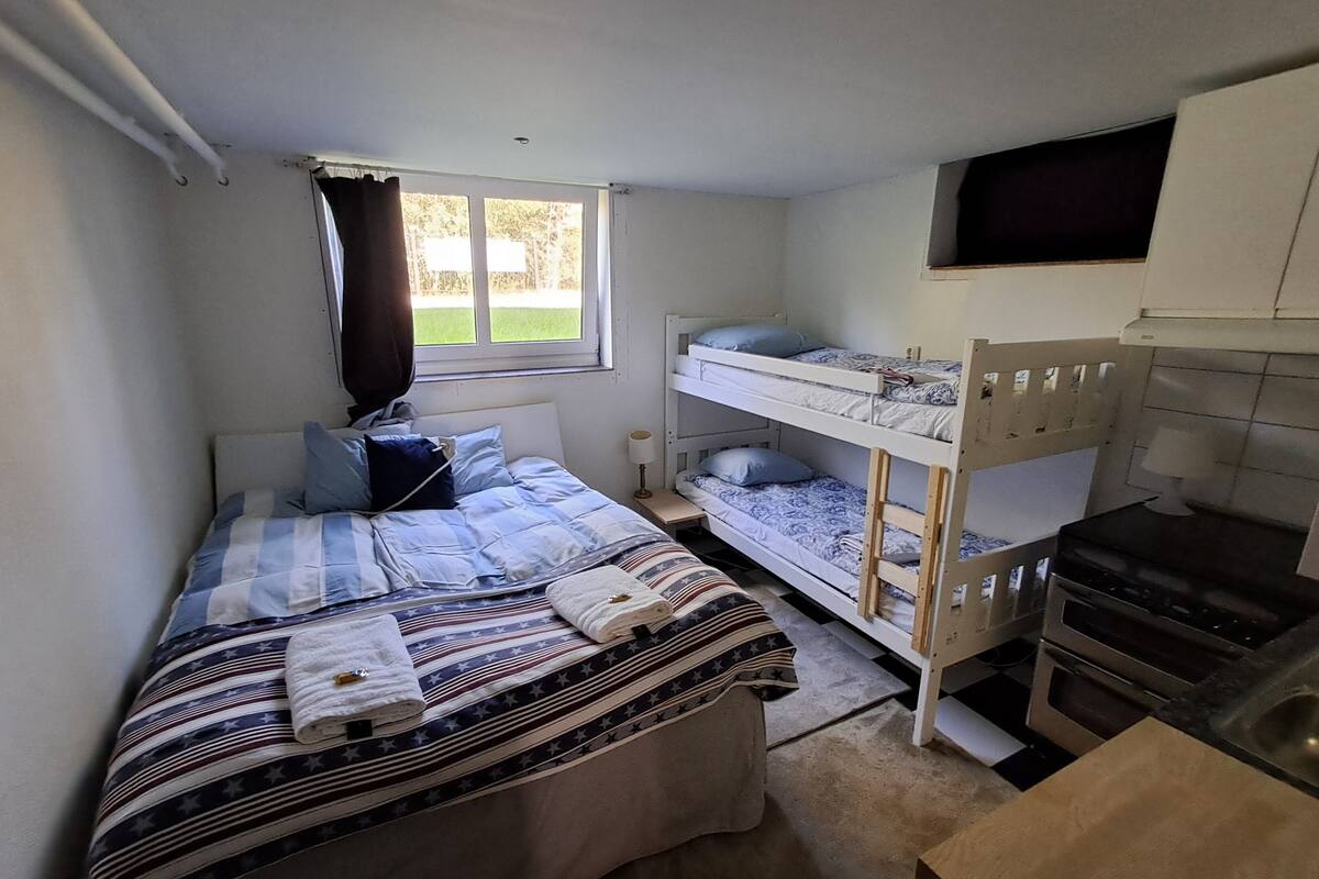 Una habitación pequeña y funcional con una cama individual, una litera, una kitchenette y una ventana con cortinas.