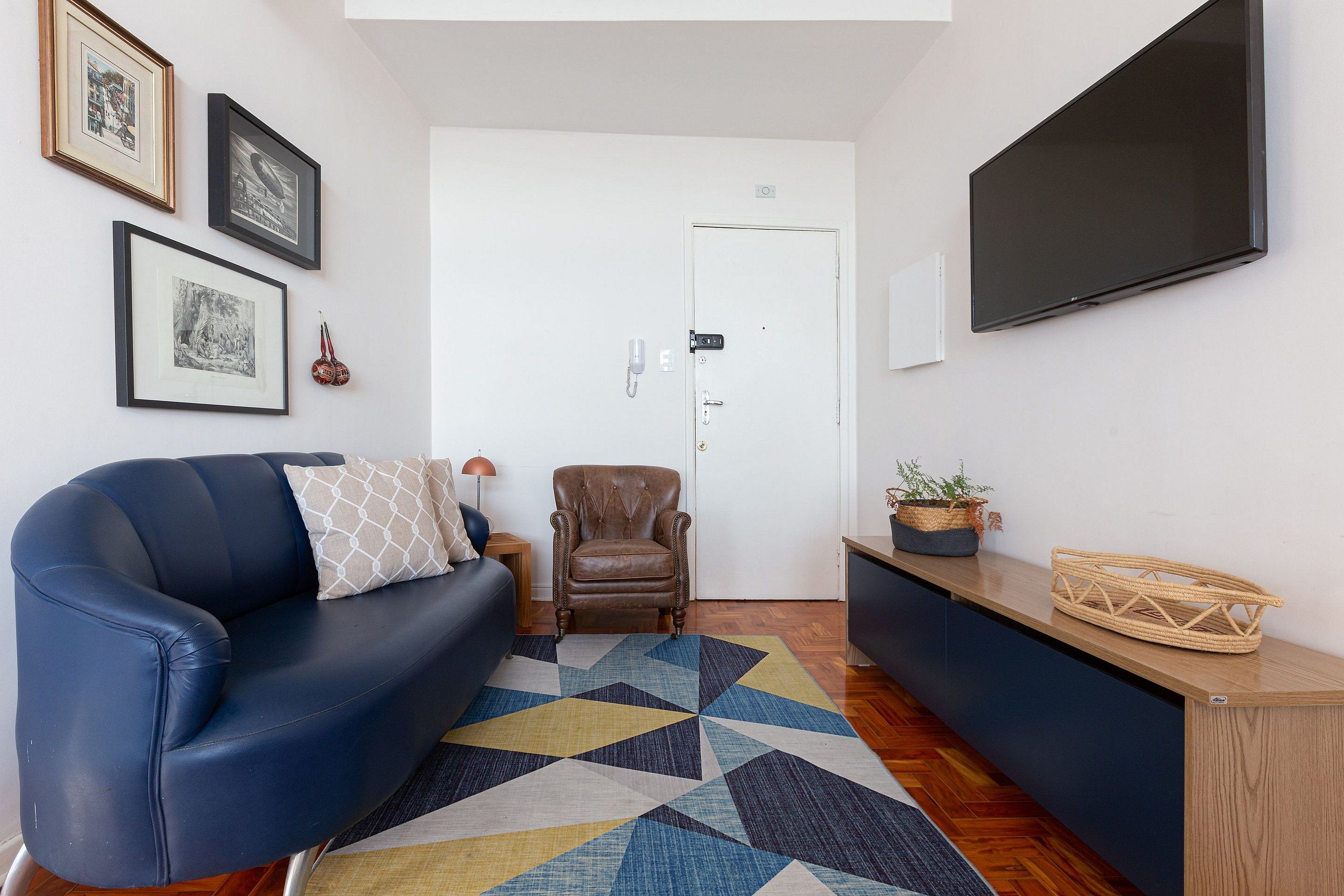 Un salon moderne avec un canapé bleu, un fauteuil en cuir, un tapis à motifs géométriques et un téléviseur à écran plat fixé au mur.