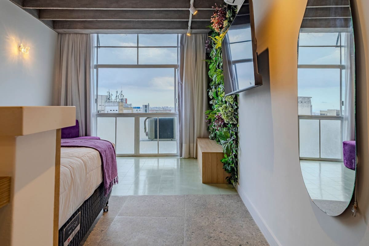 A modern hotel room with a bed, a large mirror, a TV, and a balcony with a city view.