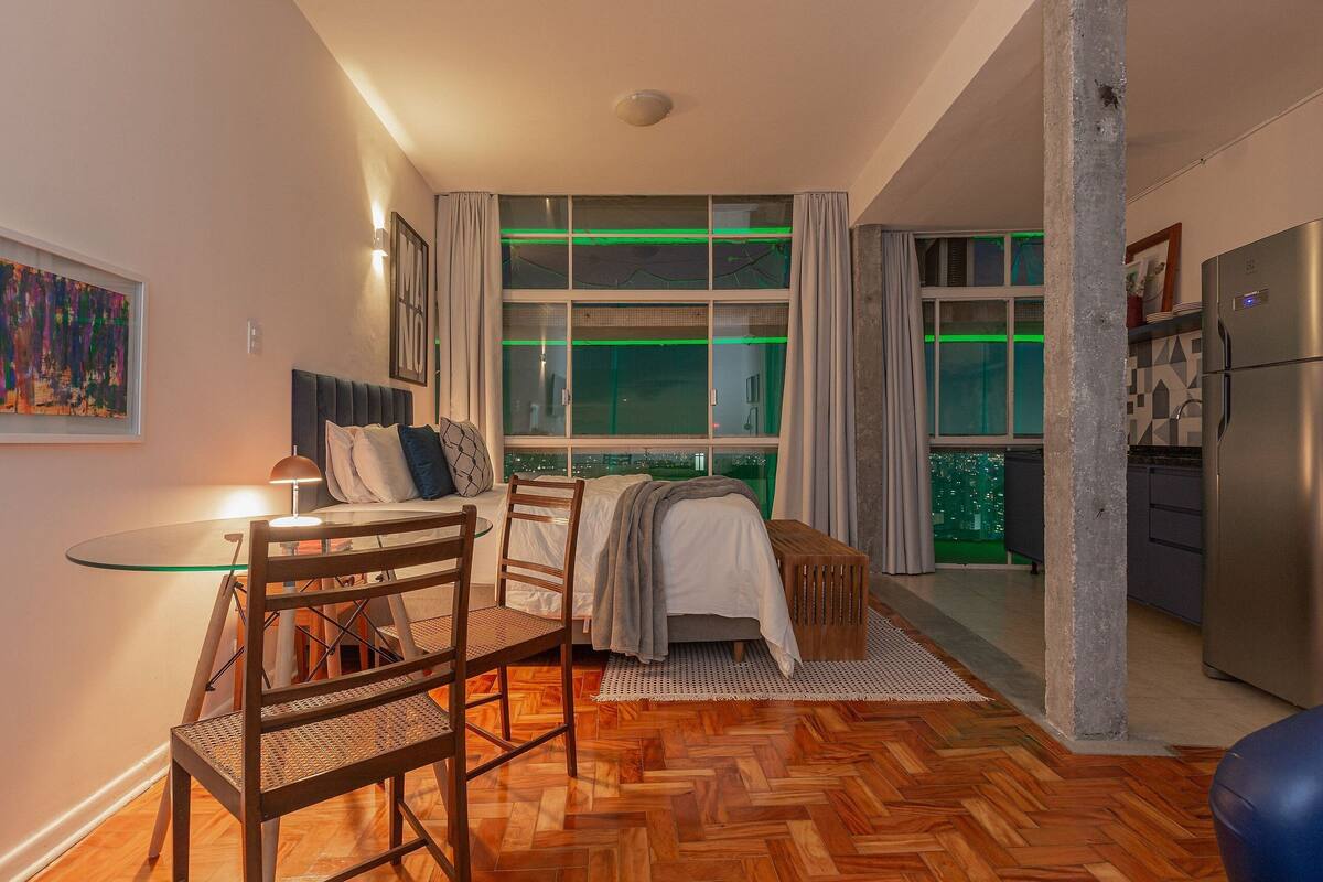 A modern bedroom with a bed, a glass table, and a chair. There is a window with curtains and a view of the city.