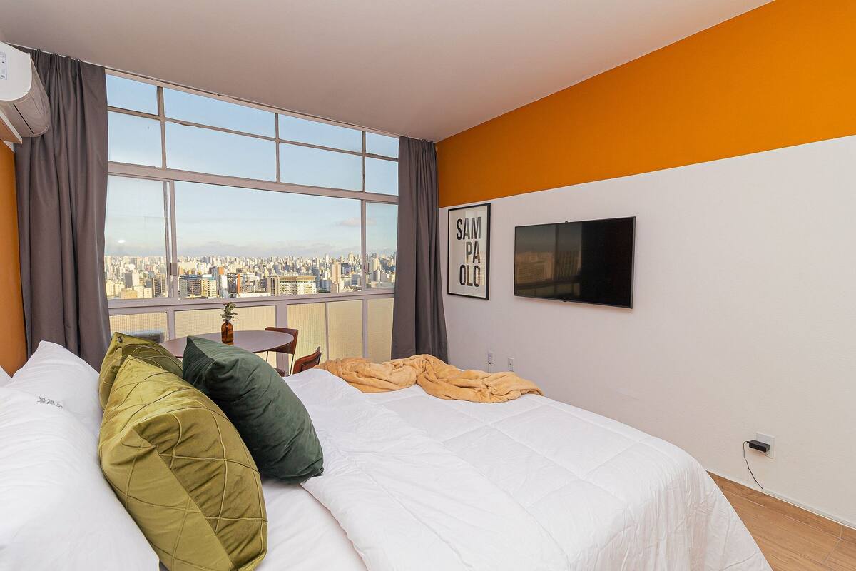 A hotel room with a large window, a bed with pillows, a TV on the wall, and a city view.