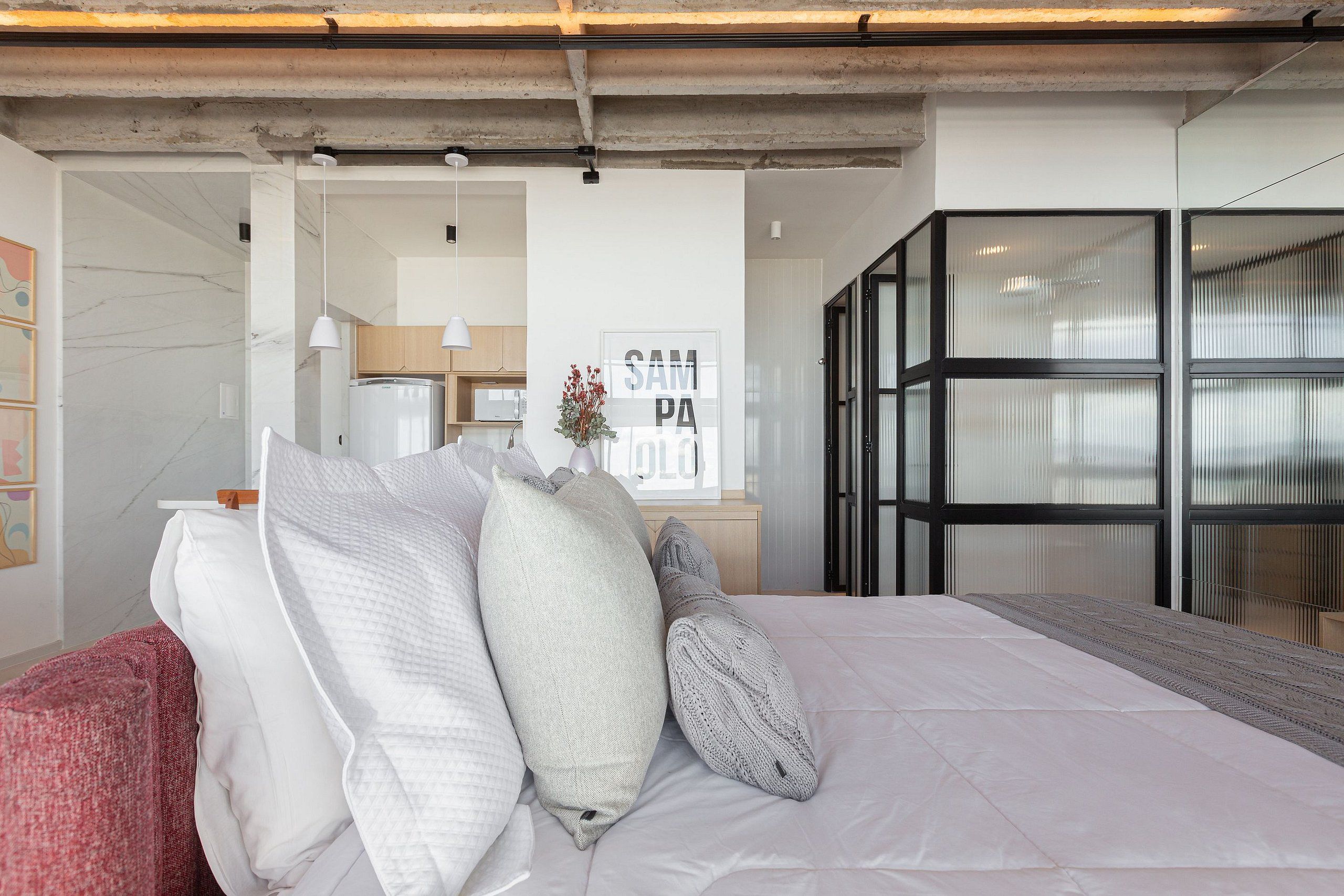A modern bedroom with a large bed, a wooden nightstand, and a view of a kitchen and living area through a glass door.