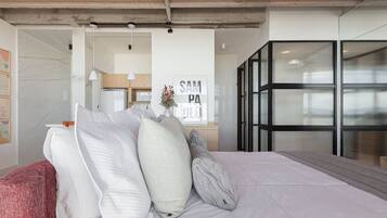 A modern bedroom with a large bed, a wooden nightstand, and a view of a kitchen and living area through a glass door.