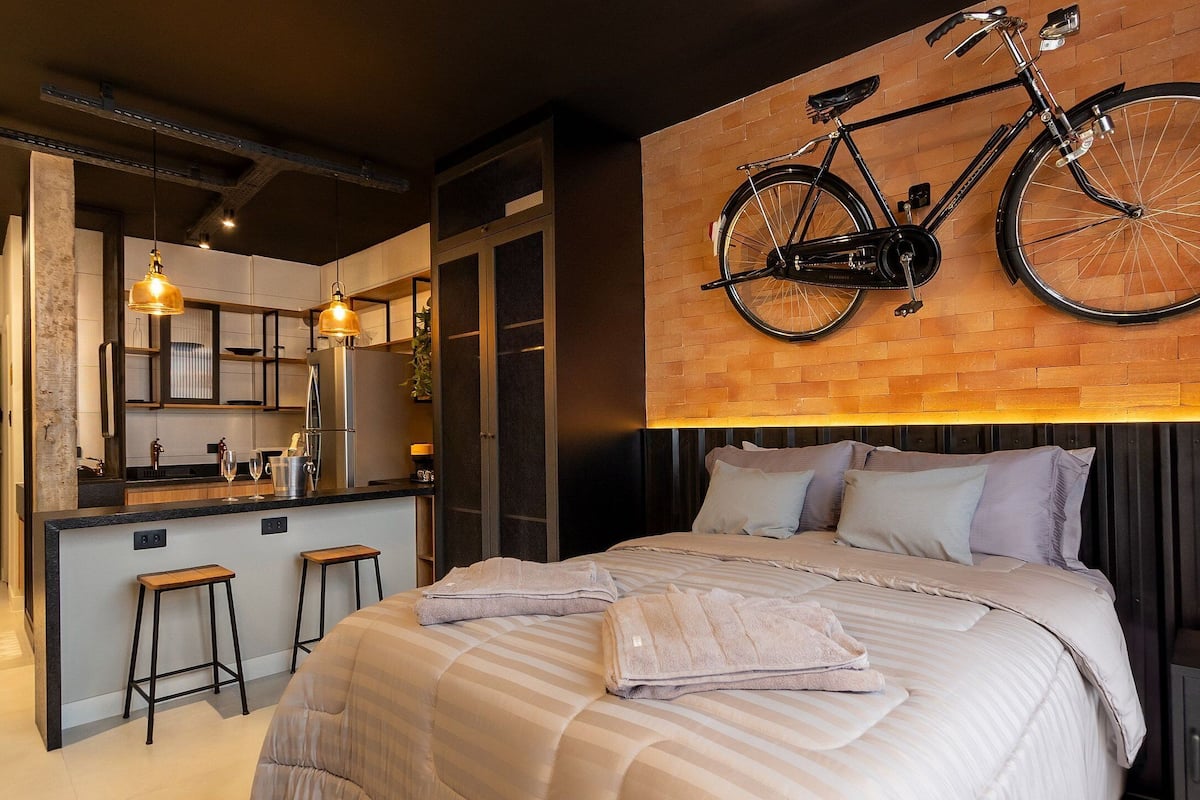 A modern bedroom with a large bed, a bicycle mounted on the wall, and a kitchen area visible through an open door.