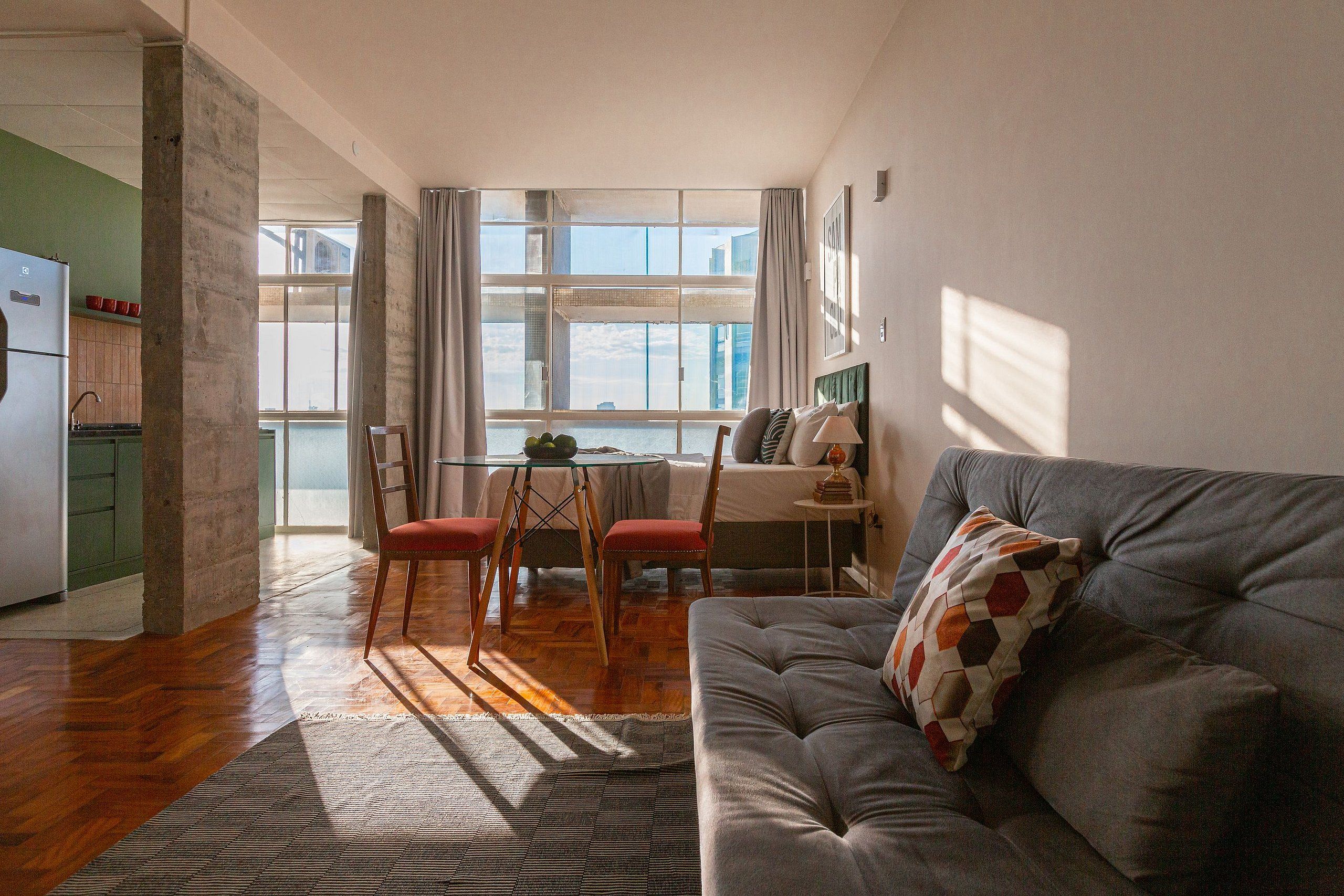 Una sala de estar moderna con un sofá gris, un comedor con mesa de vidrio, y una cocina visible a través de una puerta abierta.