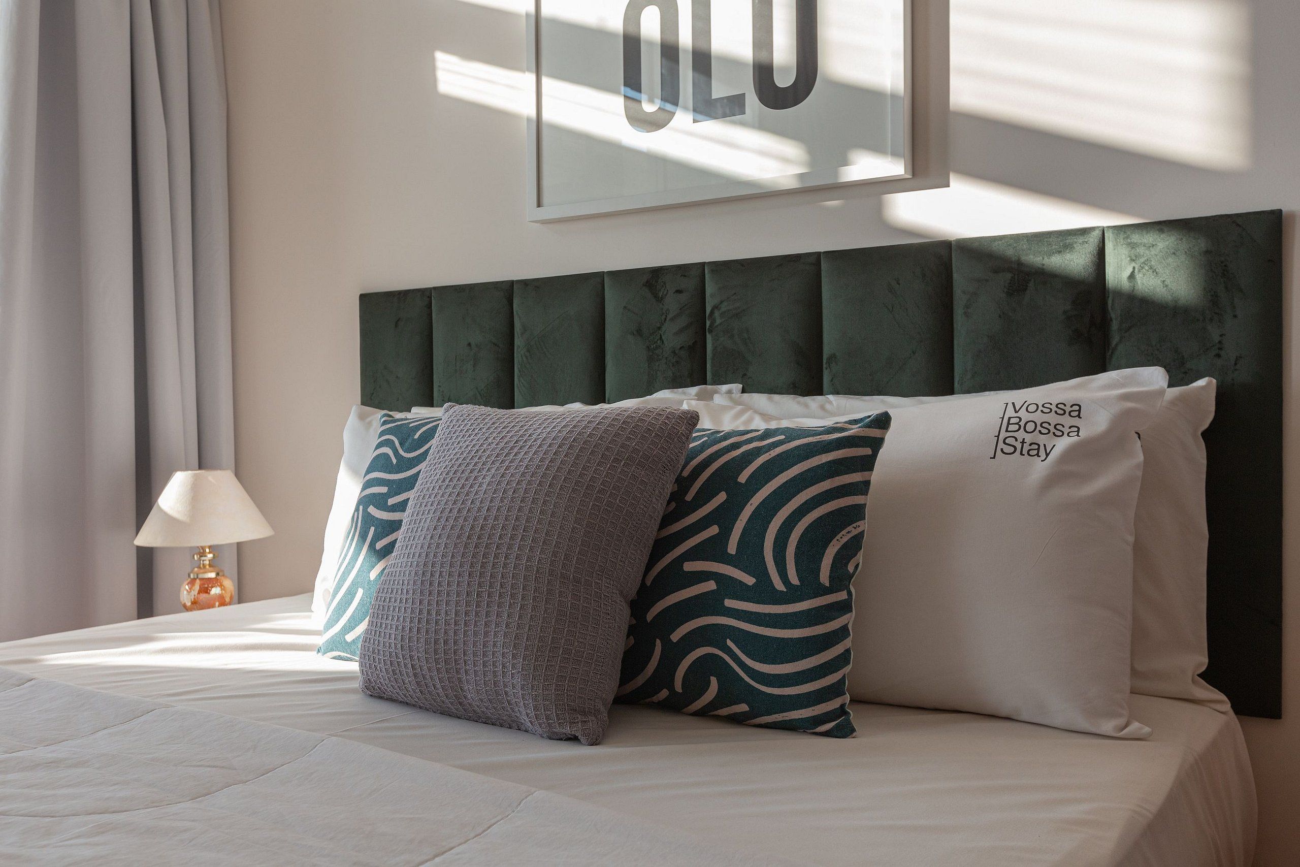 A neatly made bed with a green upholstered headboard, a lamp, and a framed sign above the bed.