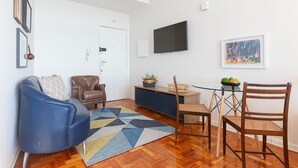 A living room with a blue sofa, a brown leather chair, a wooden dining table with chairs, a glass coffee table, and a colorful rug. - Vossa Bossa Copan (São Paulo)