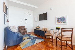 A living room with a blue sofa, a brown leather chair, a wooden dining table with chairs, a glass coffee table, and a colorful rug. - Vossa Bossa Copan (São Paulo)