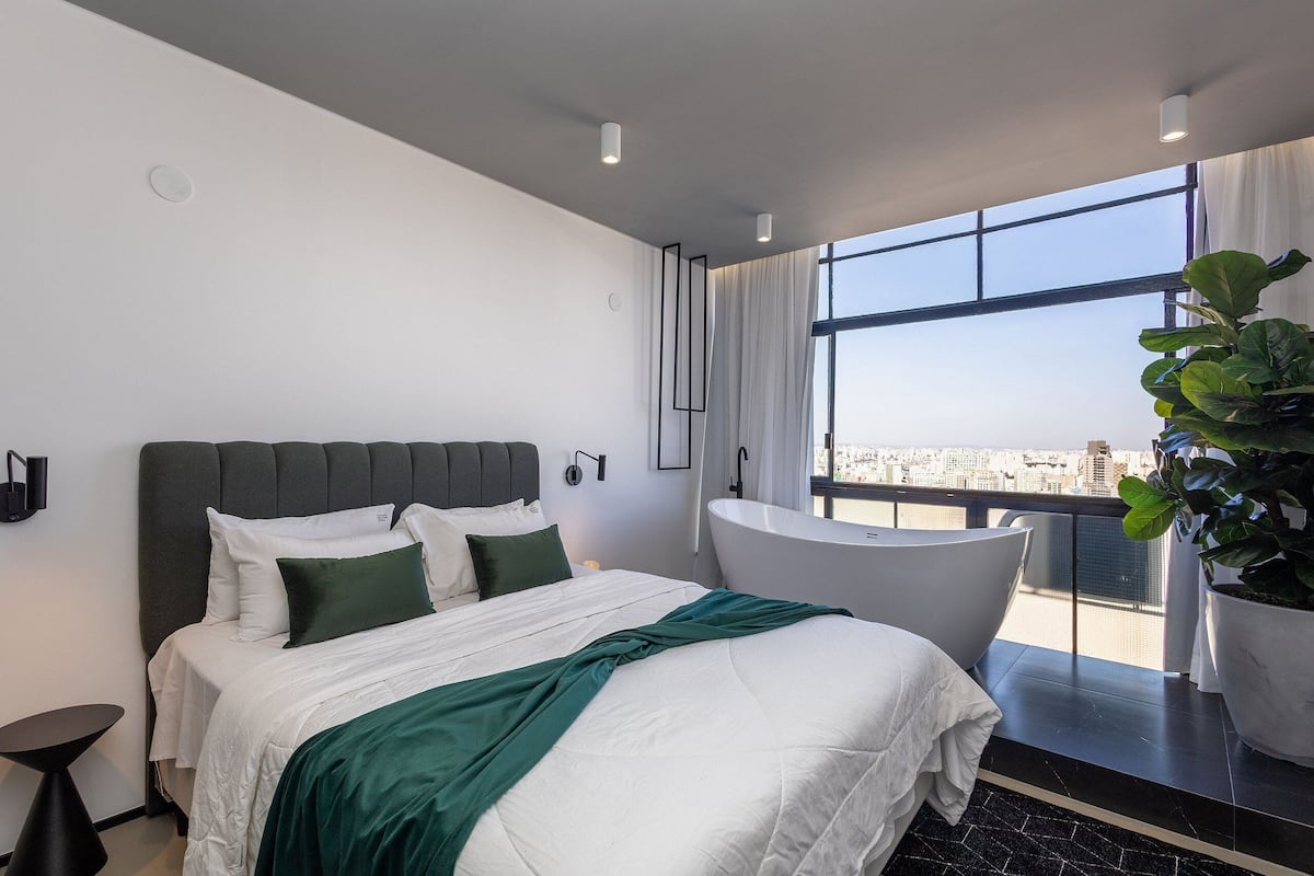 A modern hotel room with a large bed, a bathtub, and a view of the city.