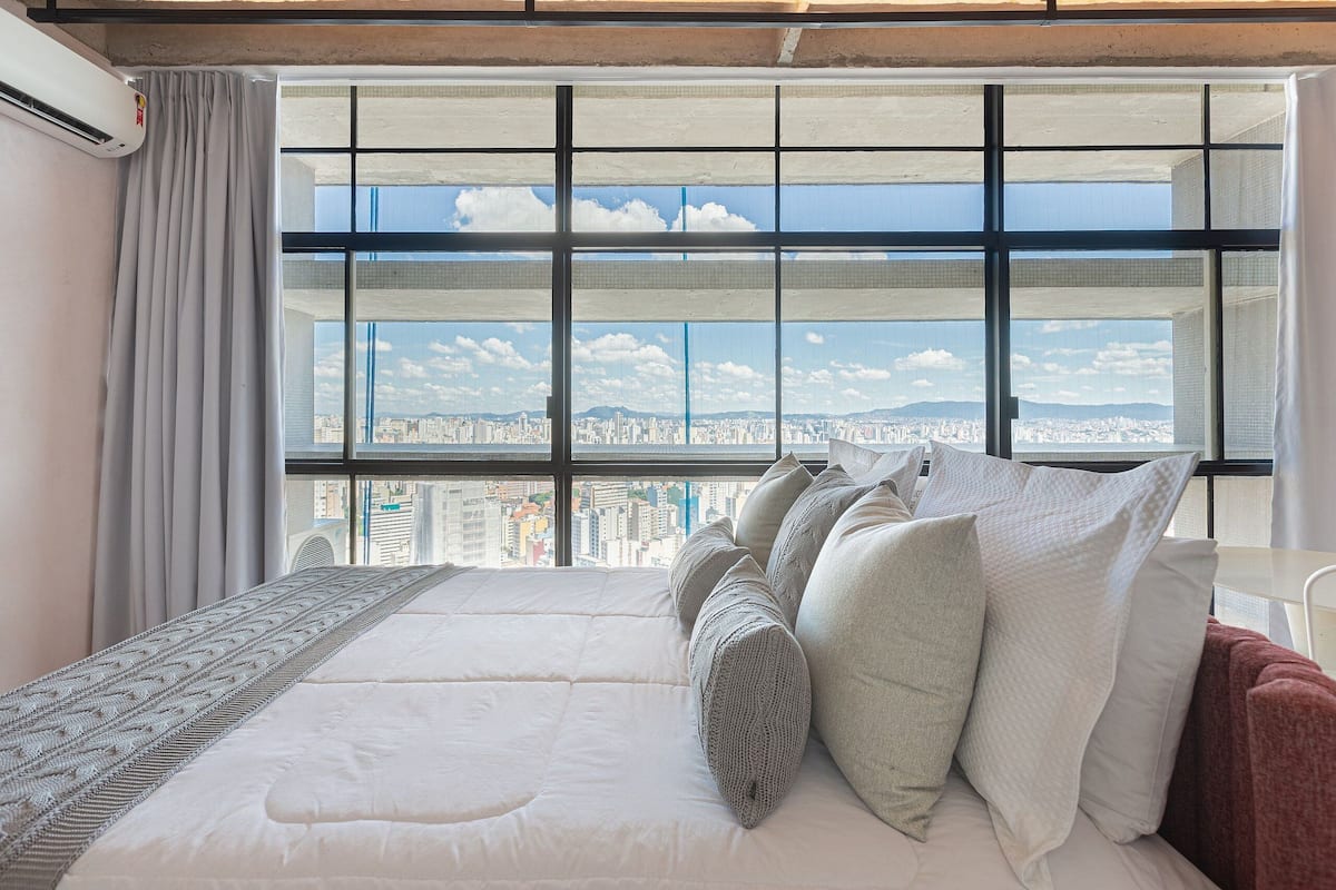 A hotel room with a large window, a bed with white bedding, and a view of a cityscape.