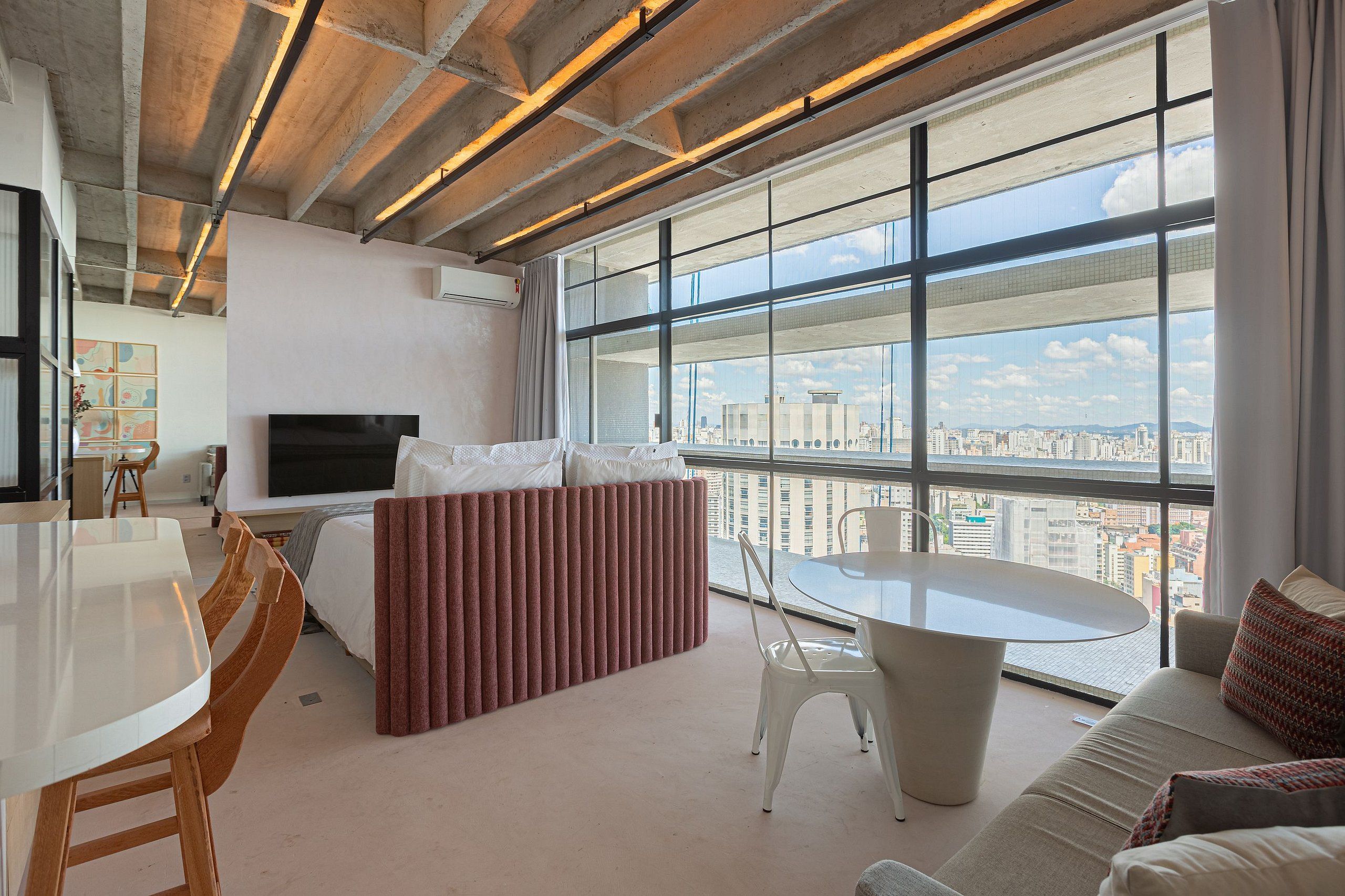 A modern hotel room with a large window, a bed, a dining area, and a sofa.