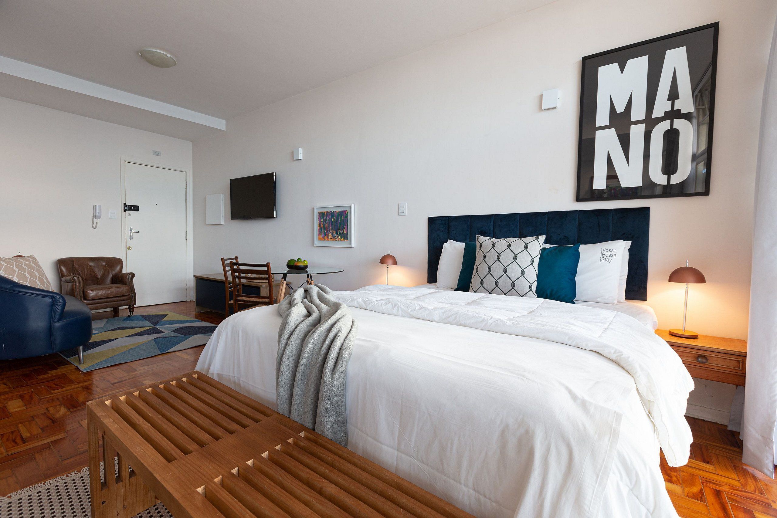 Habitación de hotel con una cama grande, un banco de madera, un escritorio con una silla, un televisor en la pared y una obra de arte enmarcada.