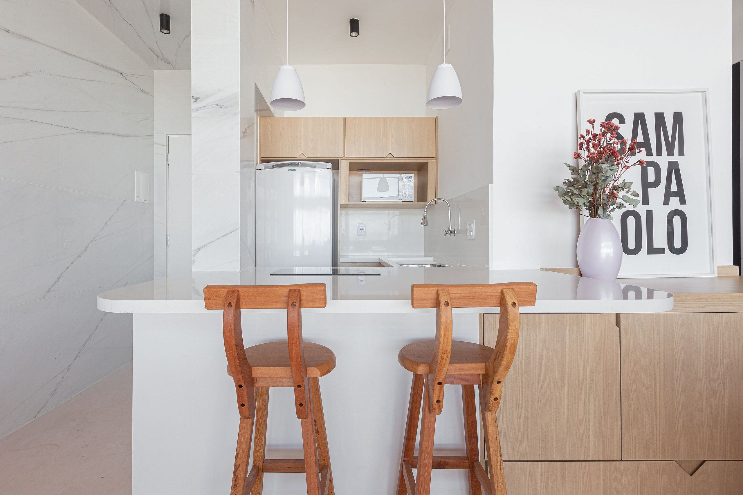 Une cuisine moderne avec des murs en marbre blanc, des armoires en bois, un îlot central blanc avec deux tabourets de bar en bois, un four à micro-ondes et un évier.