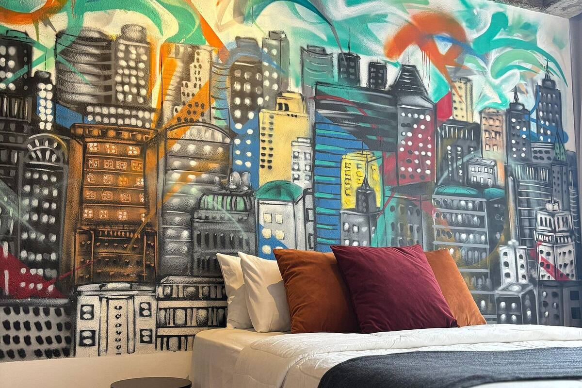 A hotel room with a bed, pillows, and a cityscape mural on the wall.