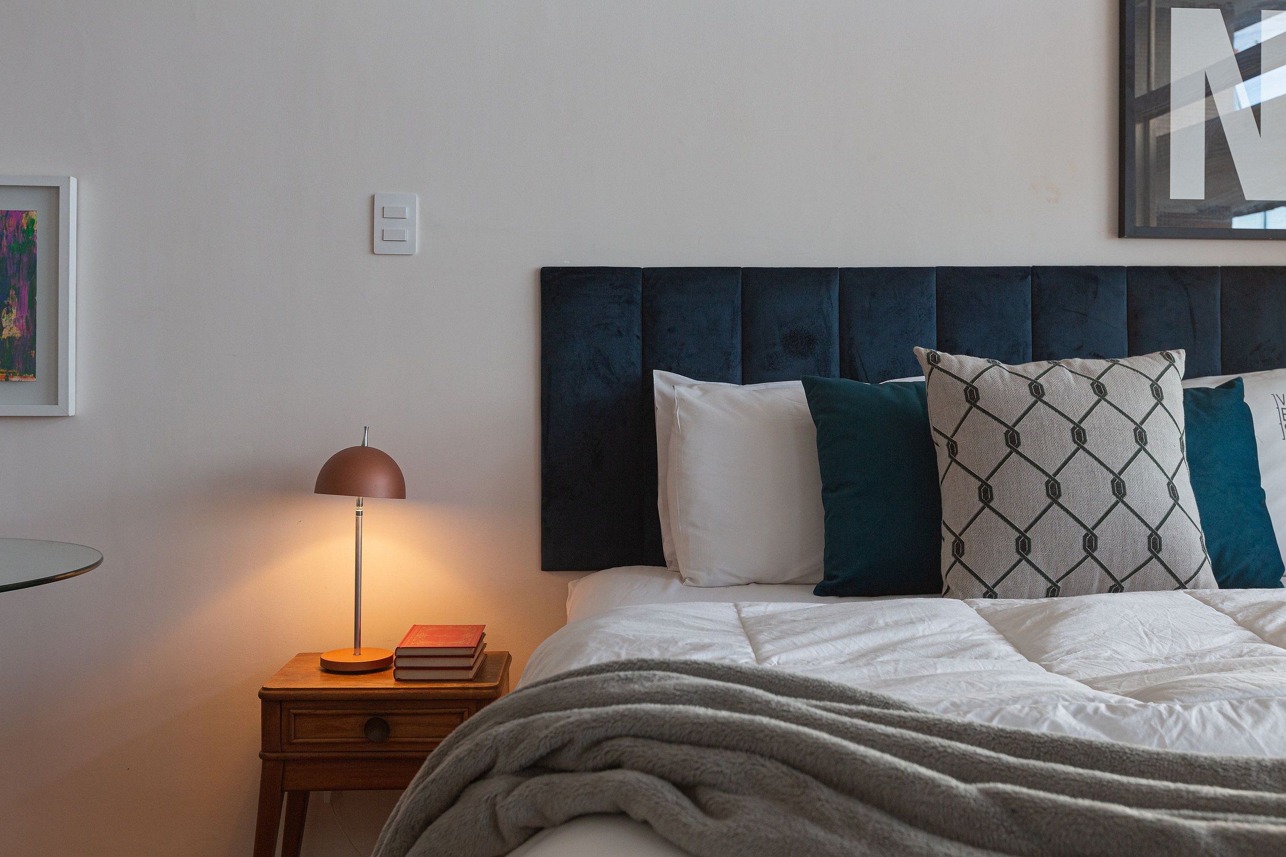 A neatly made bed with a dark headboard, a bedside lamp, and a small wooden nightstand with a book and a lamp.