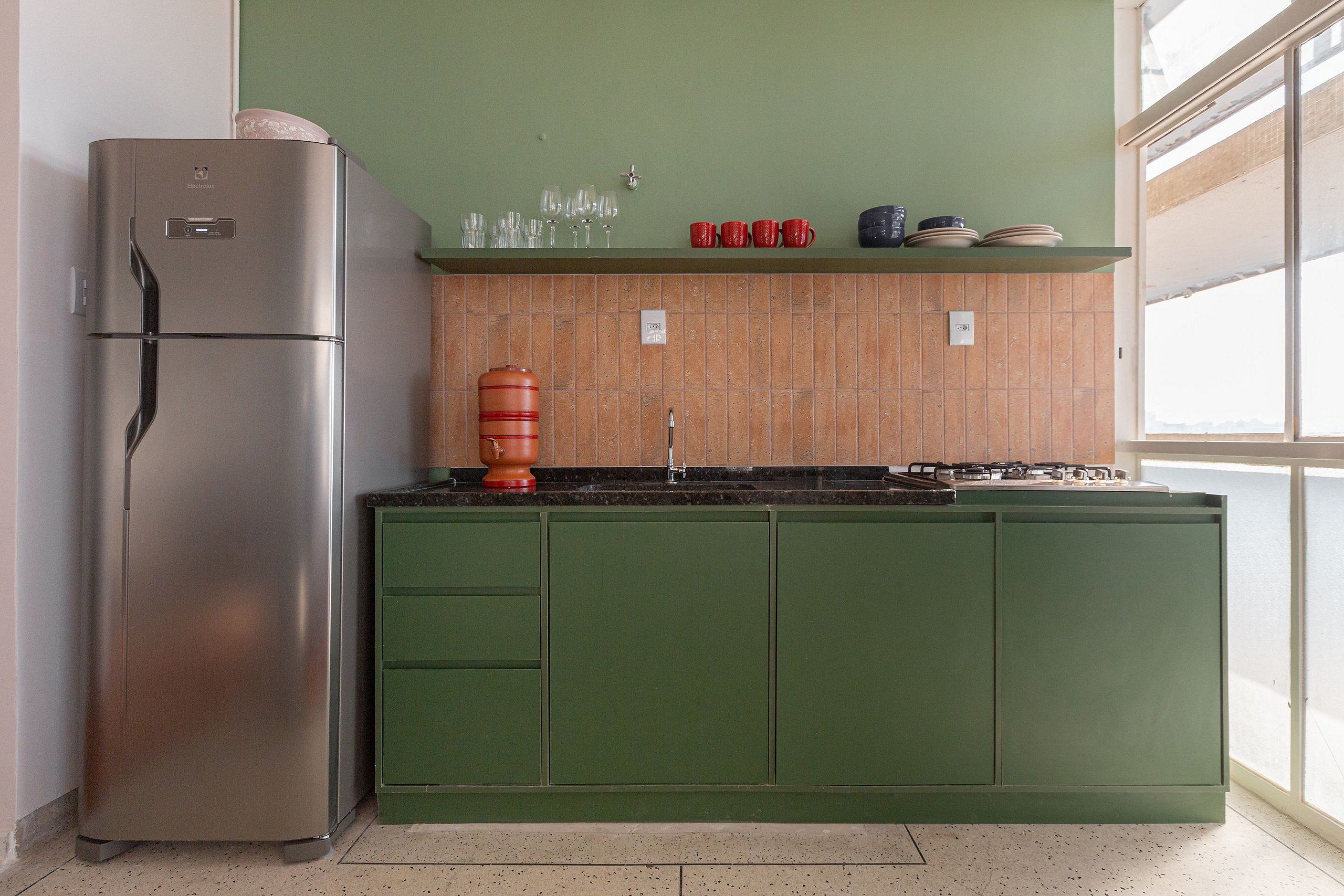 A kitchen with a stainless steel refrigerator, green cabinets, a tiled backsplash, and a window providing natural light.